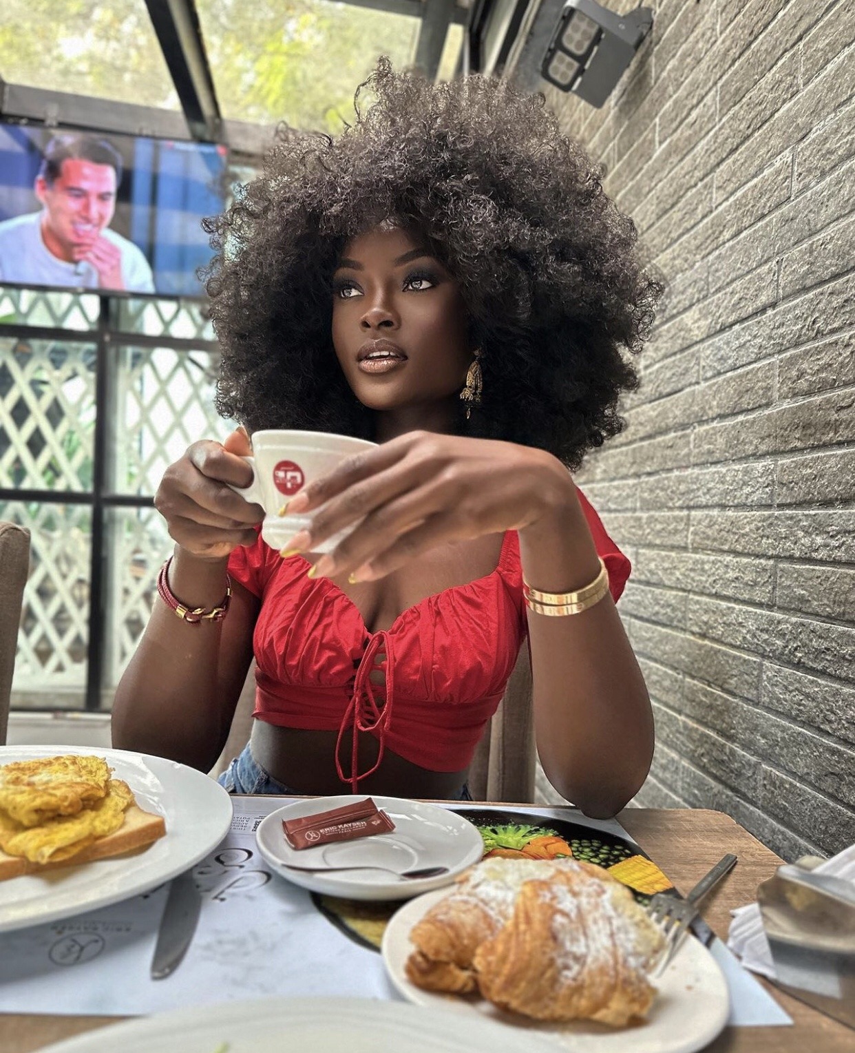 a beautiful black woman with an afro hairstyle is sitting at a table in a cafe. she's wearing a red top and holding a cup of coffee. she's looking away pensively. there are some pastries and breakfast on the table in front of her.