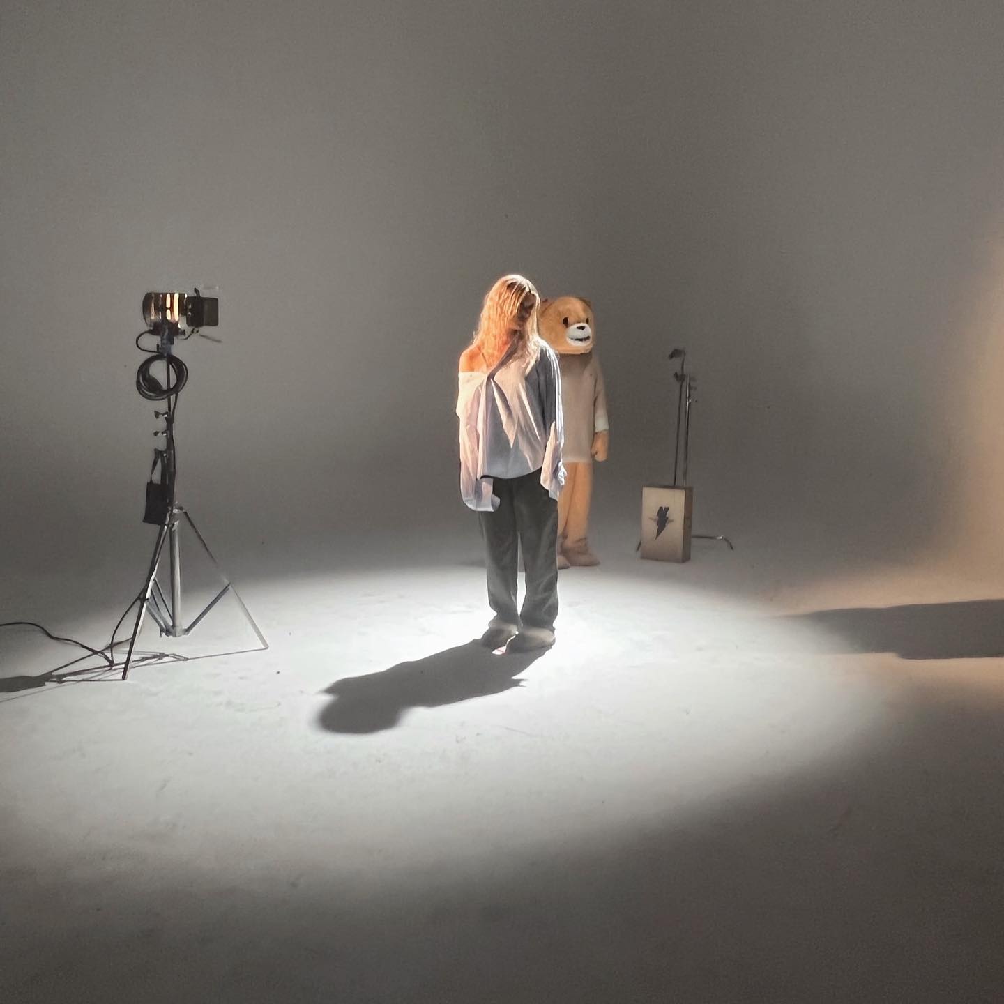 a woman stands in a studio with lighting equipment to her left and a person in a teddy bear costume behind her. a microphone stand and a box with a lightning bolt are nearby. the woman is wearing an offshoulder shirt and pants, and her shadow is cast on the grey background.