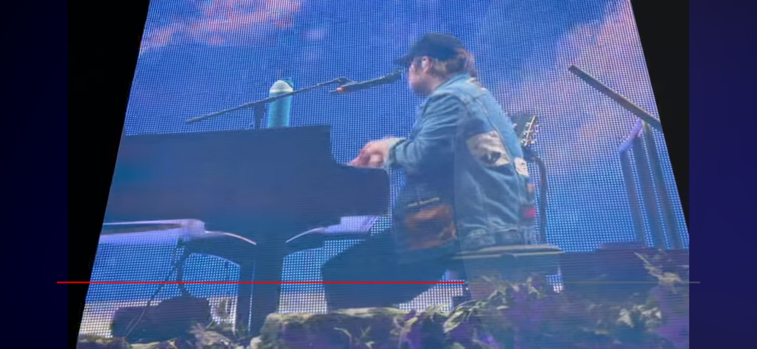 a man in a blue jean jacket and black baseball cap is sitting at a piano and singing into a microphone. he is on a stage with a guitar to his right, and a microphone and a water bottle to his left. the stage is in front of a large blue screen. there are people in the audience, but they are out of focus.  the lighting is dark and blue.