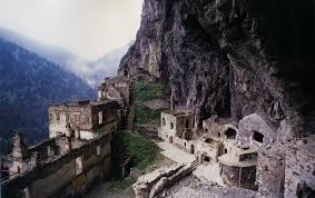 the image shows the sumela monastery clinging to a cliffside in turkey, set against a mountainous landscape with a cloudy sky. the monastery consists of stone buildings, partially in ruins, built into the rock face. the architecture is historical, and the site is a tourist attraction, showcasing a blend of ancient architecture and natural scenery.