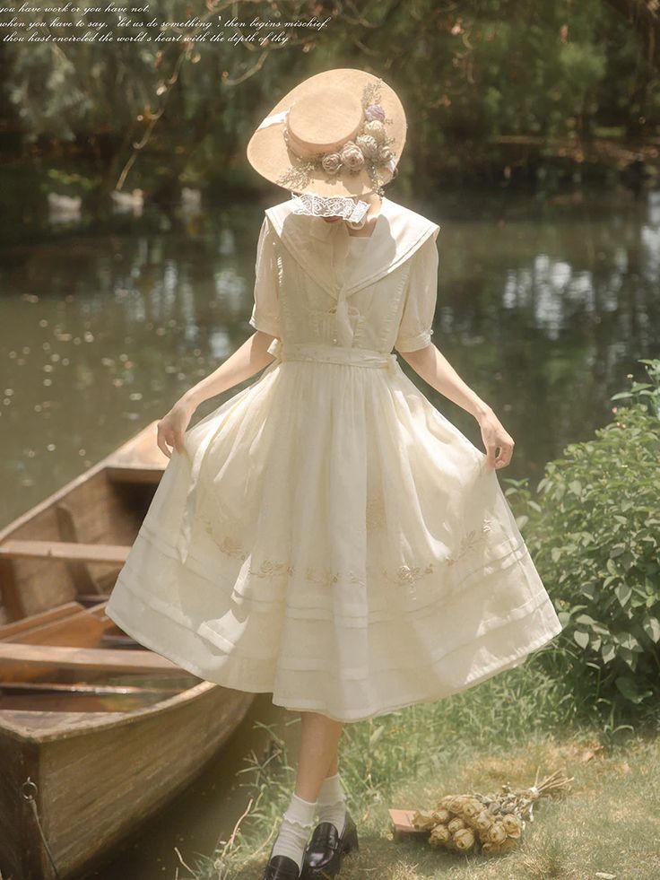 a young woman in a vintageinspired cream dress and straw hat stands by a rustic row boat near a river. the dress is flowy with lace details and a sailor collar. she's holding out the skirt slightly with both hands, and her head is tilted down, obscuring her face. the setting is idyllic and natural, with sunlight filtering through the trees in the background.  she has a bouquet of dried flowers laying on the ground by her feet.