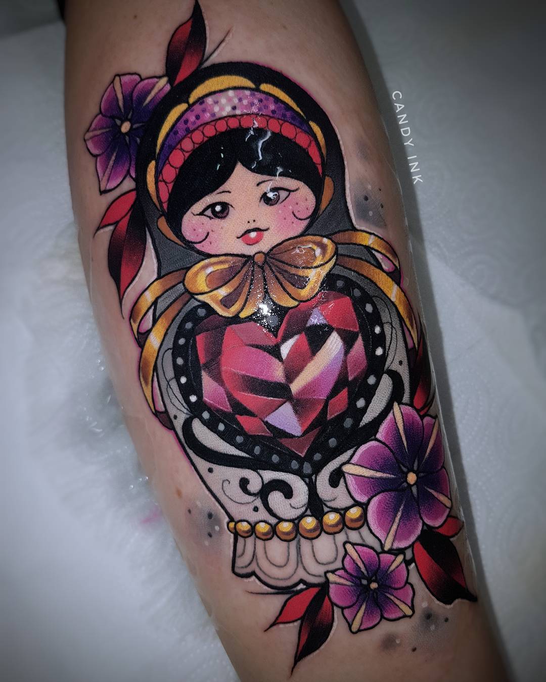 this is a photograph showcasing a vibrant neotraditional tattoo of a matryoshka doll, also known as a russian nesting doll. the doll's attire is rendered in a striking color palette of deep purples, rich golds, and fiery reds. she prominently displays a heartshaped gem on her chest, further emphasizing the theme of love and affection often associated with these dolls. the tattoo is beautifully framed by delicate purple flowers with green leaves, adding to the overall aesthetic appeal.  the artist's signature, "candy ink," is subtly placed alongside the tattoo.