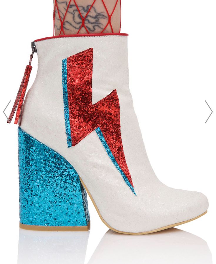 a closeup shot showcases a pair of women's white glitter ankle boots. the boots feature a chunky block heel with a teal glitter accent, and a red and teal glitter lightning bolt design on the side.  the boots are shown on a white background with a subtle reflection below. a woman's leg, wearing red fishnet stockings, is visible at the top of the image.  the image is presented as if on a product page, with navigation arrows to the left and right.