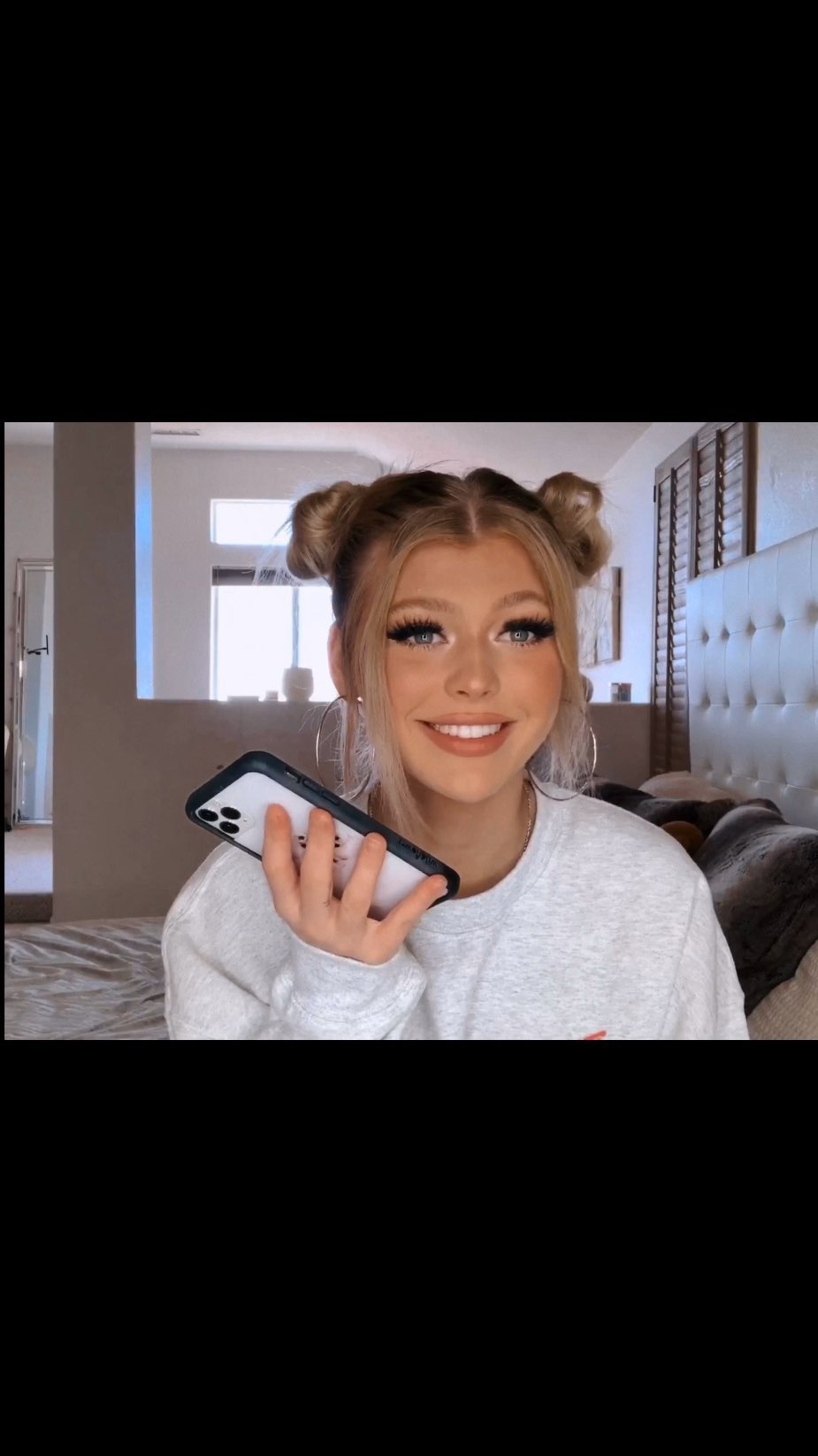 a smiling woman with a bun hairstyle is taking a selfie while holding her phone. she's wearing earrings and a gray sweatshirt. she appears to be in a bedroom.