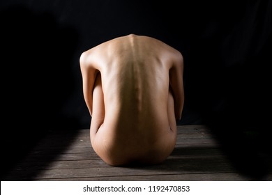 a nude woman is crouching on a wooden floor with her back to the camera. her spine is visible and her skin is pale. the background is dark and there are shadows on the floor.