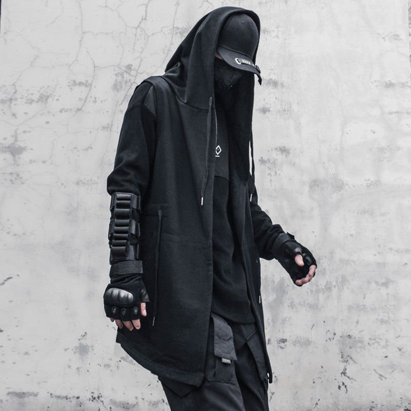 a stylish man in all black clothing poses for a fashion shoot. he is wearing a black hooded jacket, black pants, and black gloves. he looks cool and mysterious. the background is a plain grey wall. the overall vibe of the photo is dark and edgy.