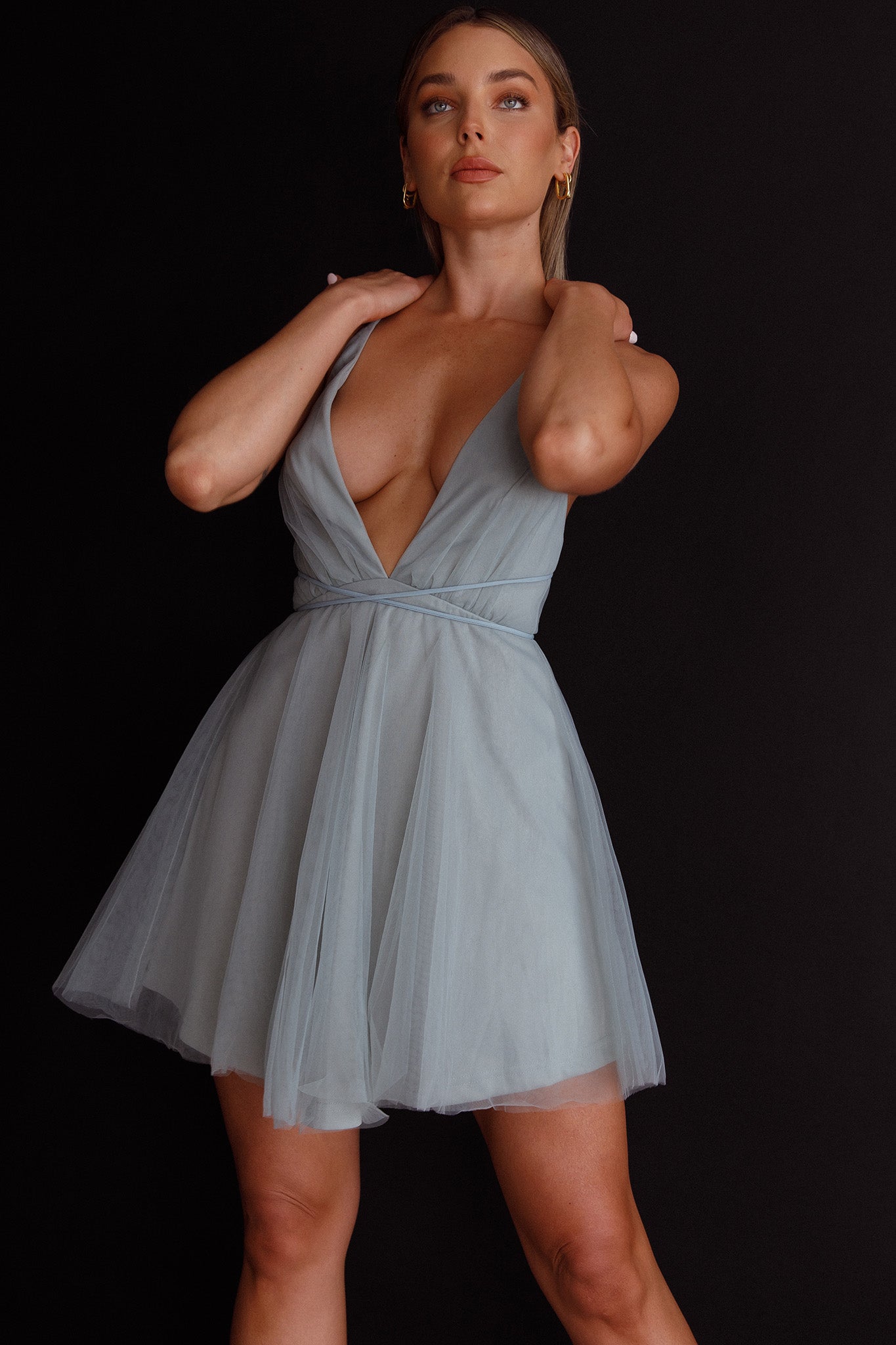 a blonde woman in a light blue tulle cocktail dress poses against a black background. the dress has a deep vneckline and a flowing skirt. she is wearing her hair down in loose waves. the lighting is soft and flattering, highlighting the model's features and the texture of the dress.