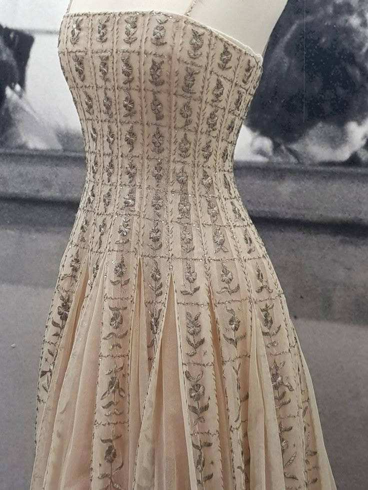closeup shot of a strapless, beaded, and embellished gown. the dress has a fitted bodice and a tulle skirt. the material has a textured appearance with intricate patterns. the dress appears to be a light beige color and is sleeveless. the background is blurred, but it appears to be taken in a museum or exhibit setting. the overall impression is one of vintage elegance and formal wear.