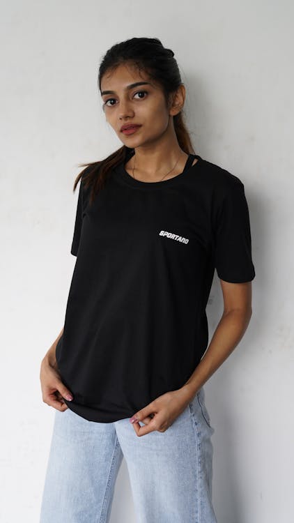 a fairskinned woman is posing indoors wearing a black tshirt with the word "sportano" printed in white on the left side of the chest. she is also wearing light wash, wideleg jeans. the background is lightcolored and plain. she has long dark hair pulled back in a low ponytail. her hands are visible and she is smiling slightly. the shot appears to be taken in a studio.