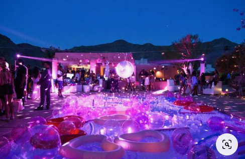 a lively pool party taking place at night with a crowd of people enjoying themselves, dancing and socializing. the environment is outdoors with mountains visible in the background, suggesting a desert resort setting. the lighting is vibrant and colorful with purple hues dominating, creating a festive and energetic atmosphere. there's a disco ball hanging above the pool, inflatable toys and floats are scattered throughout the pool, all illuminated by bright spotlights.