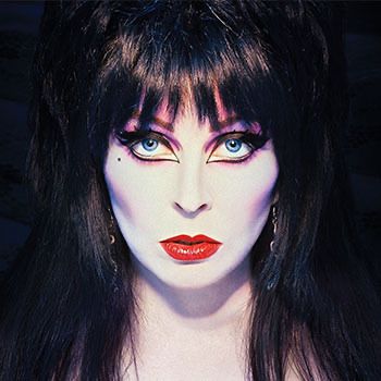 he image shows a woman with long black hair, blue eyes, and red lipstick. she is wearing a costume that is characteristic of a witch. her makeup is dramatic, with very dark eyeliner and eyeshadow. she is looking directly at the camera. the image appears to be from a movie or television show.
