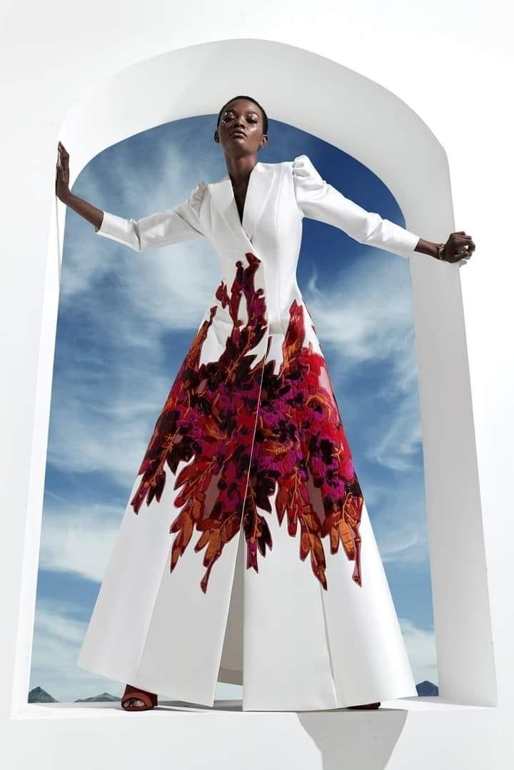 a black woman wearing a white dress with red flowers is posing in front of a white arched background. she has her arms outstretched, and the dress is flowing around her. the dress is long and has a split in the front. the woman is wearing red shoes, and the sky is visible in the background.