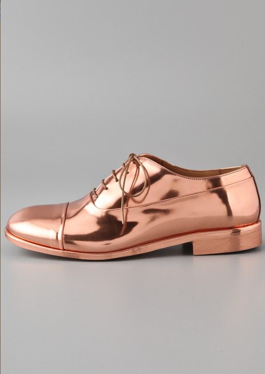 the image showcases a single rose gold metallic oxford shoe. the shoe is a women's style, featuring a shiny, coppercolored finish and classic laceup detailing.  it has a low heel and a slightly pointed toe. the shoe is displayed against a neutral gray background, allowing the metallic finish to be the focal point. the image appears to be a product shot, likely for an online retailer or fashion magazine.