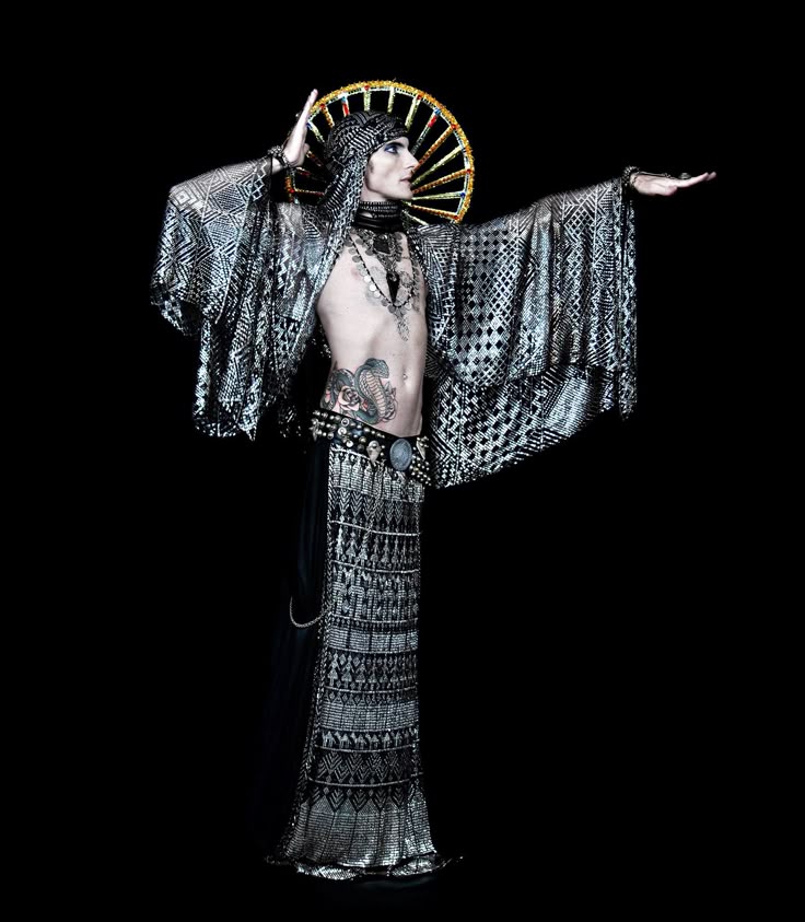 a person stands in a fulllength shot against a black backdrop, exuding a sense of egyptianinspired fashion. the individual is dressed in an open robe, a long skirt, and a headdress, all featuring geometric patterns. a halo adorns the headdress, adding to the mystical aesthetic. a belt with studded detailing cinches the robe, accentuating the waist. a striking cobra tattoo is visible on the abdomen, complemented by the various necklaces and jewelry embellishing the figure.