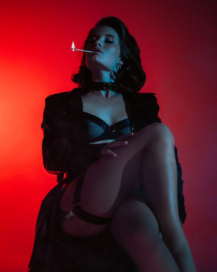 a lowangle, closeup shot of a woman with dark hair styled in a retro fashion. she is wearing a black leather jacket, black lingerie including a bralette and stockings with garter belt, and a choker necklace. she is holding a lit match near her lips in a suggestive pose against a vibrant red background, creating a strong, moody contrast. the lighting is dramatic and highlights her features and clothing, creating a film noir or vintage pinup aesthetic. her expression is somewhat aloof and mysterious. the overall mood is one of alluring confidence and sensual mystery.