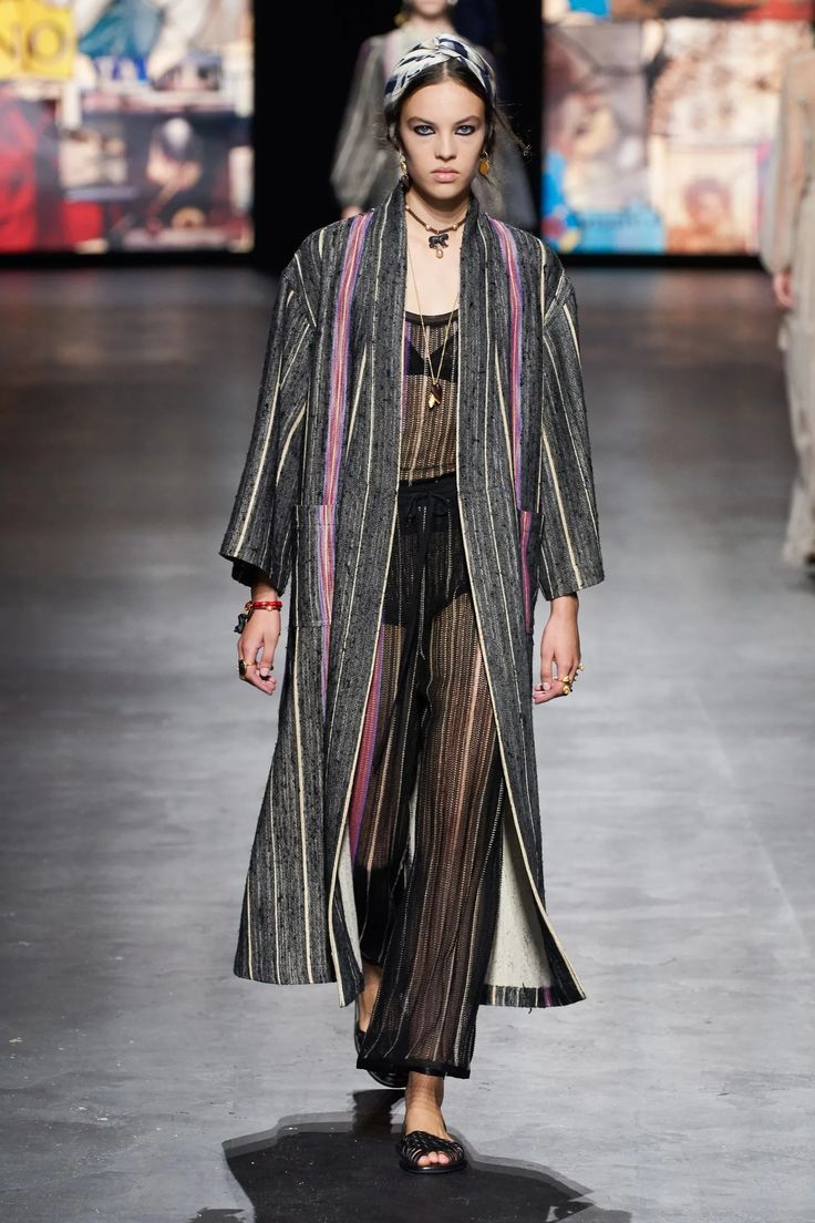 a model walks the runway in a black and white striped longsleeved coat, black pants with a sheer overlay and black flat sandals. she is wearing a black necklace and a headscarf. the backdrop is a runway with a blurred background.  she is looking forward with a serious expression on her face.