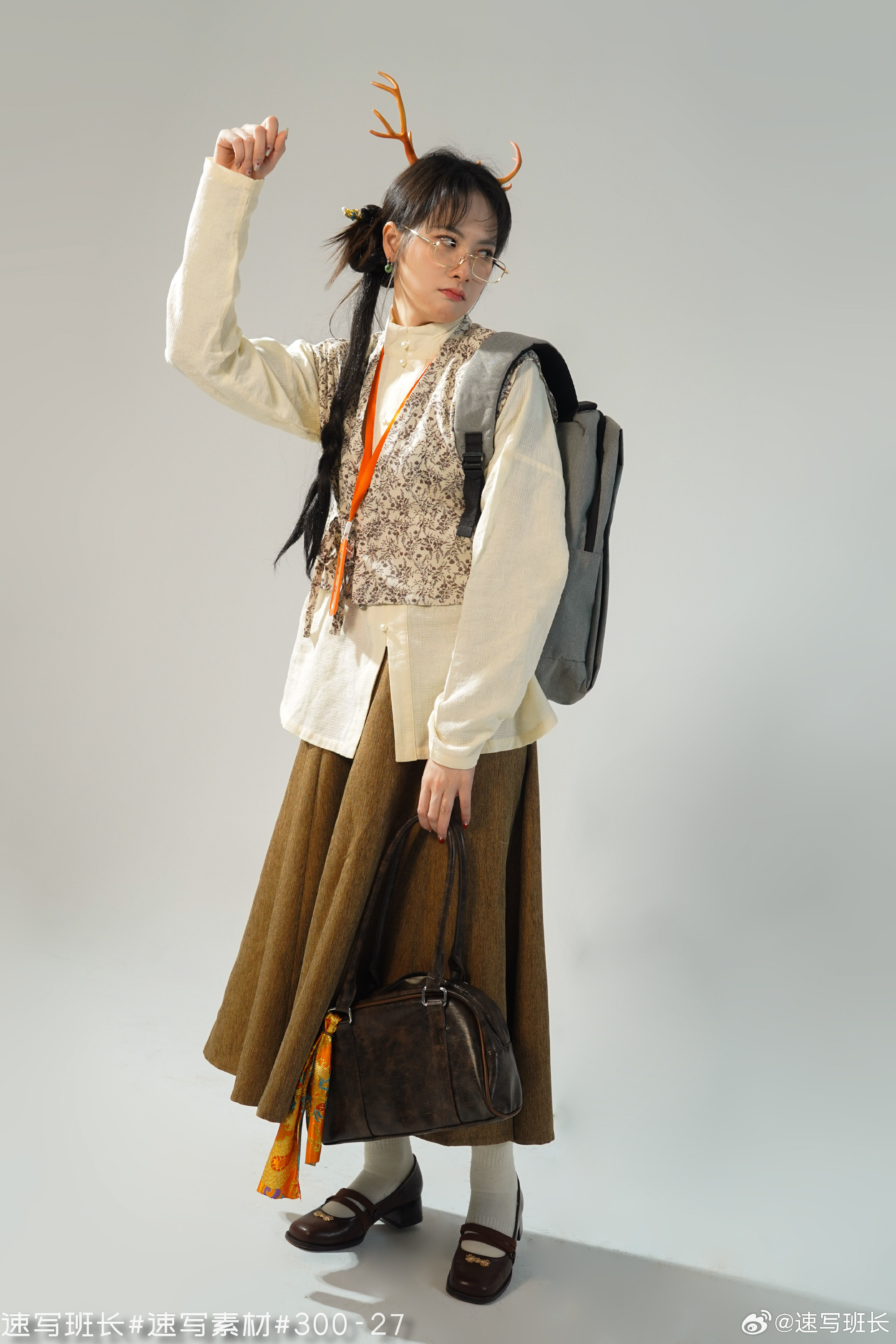 a young woman stands against a plain gray backdrop, illuminated by bright studio lighting. she is wearing a whimsical outfit, featuring a white shirt, a patterned vest, a long brown skirt, and brown shoes. she accessorizes with a backpack, a handbag, a colorful scarf, deer antlers headband, and round glasses. her pose is playful and confident.