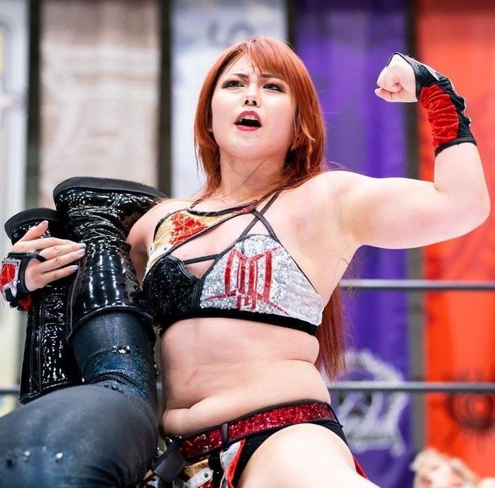 a closeup shot of a female professional wrestler with vibrant red hair. she's in the middle of a match, holding one of her opponent's legs while flexing her bicep. her wrestling attire is a striking mix of black, red, and silver, and she looks determined and confident. the background is blurred but shows the wrestling ring and some of the audience.