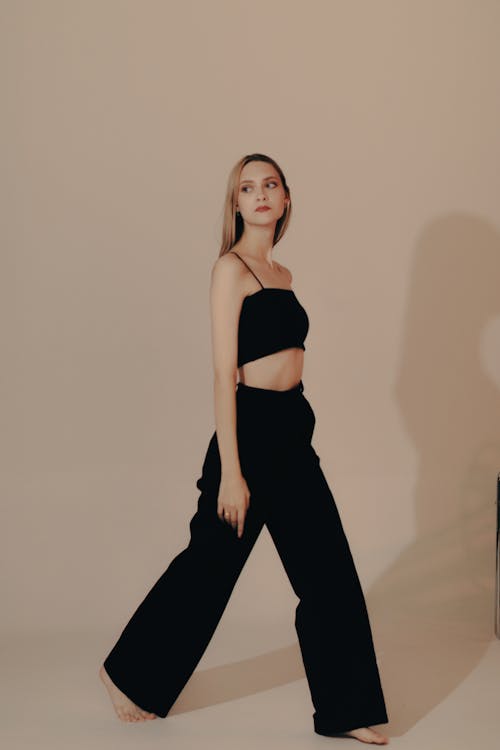 the image shows a slender, tall woman with long blonde hair walking in a studio setting. she's wearing a simple, black cropped top and wideleg black trousers. the background is a clean, light beige color, which draws focus to the model and her outfit.  the overall aesthetic is minimalist and elegant. the image is professionally lit and captured, typical of fashion photography.