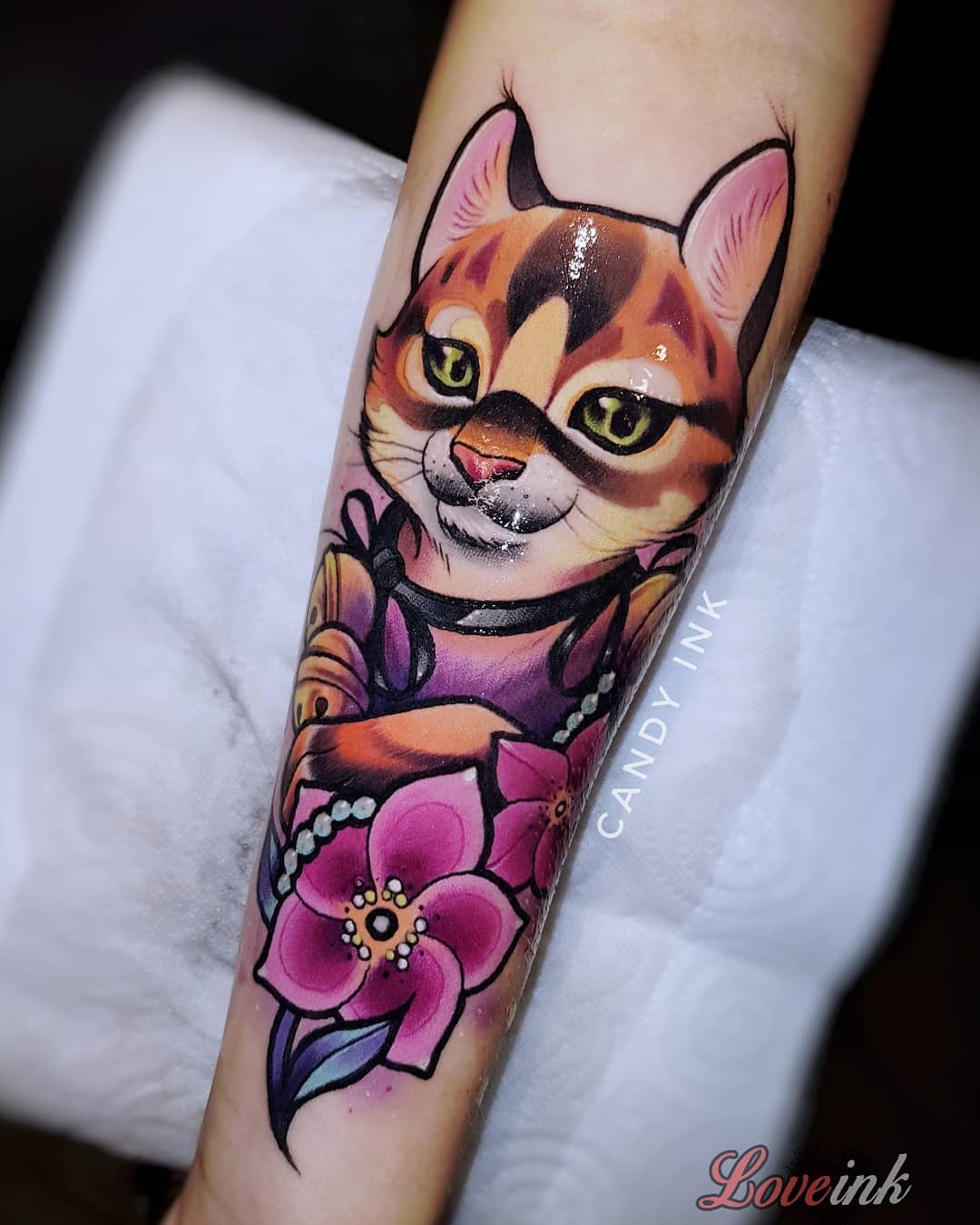 a tattoo of a cartoonstyle cat wearing a pearl necklace and surrounded by pink flowers. the tattoo is on a person's forearm. the tattoo artist's signature, "candy ink," is visible in black ink on the inner forearm. the watermark for the tattoo shop, "loveink," is visible in pink cursive at the bottom right of the image.