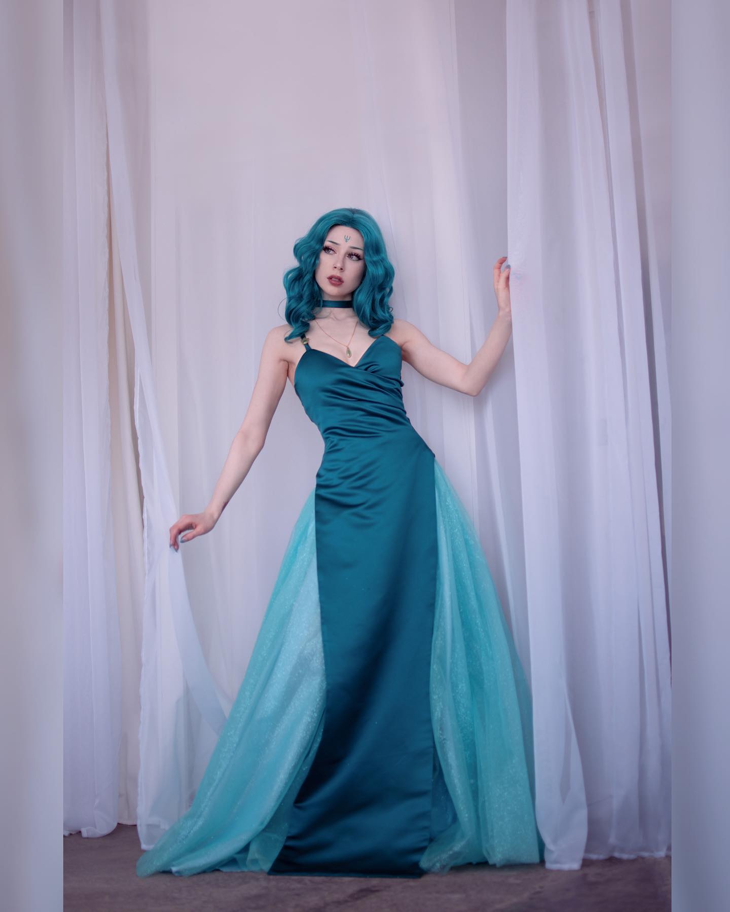 the image is a fullbody studio shot of a woman modeling a teal and turquoise gown.  the dress is primarily a teal satin material, with a tulle overlay in a lighter turquoise color that forms a train. the woman has a teal wig styled in loose curls and is wearing a delicate choker necklace. she's posed gracefully against a backdrop of white curtains, creating a soft, ethereal atmosphere. the overall style evokes a sense of fantasy or perhaps cosplay, with the styling and pose suggesting a character from a fantasy setting or anime. the lighting and composition emphasize the elegance and detail of the dress and the model's pose.