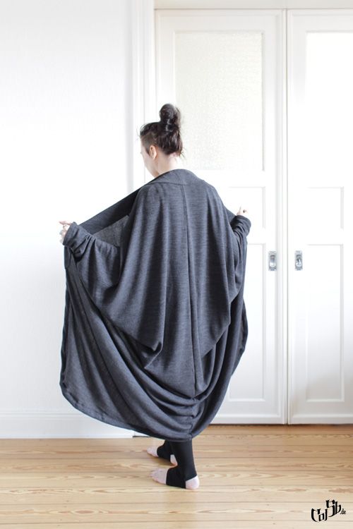 a woman with a bun is wearing a long, gray coat with a draped silhouette. she is standing in front of a white door with a wooden floor beneath her feet. the coat appears to be a stylish and comfortable fashion piece.