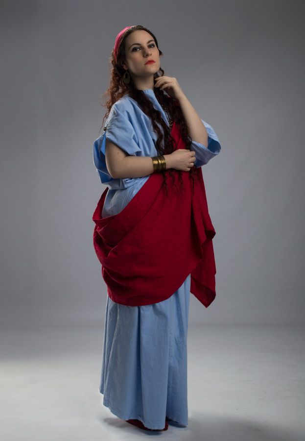 a fulllength studio shot of a woman posing in a historically accurate recreation of a roman stola and palla.  she is wearing a light blue linen tunicstyle dress underneath a crimson red palla (shawl) draped over her shoulders. she has long, dark curly hair, a pink headband, and several bronze bracelets on her left wrist. her expression is confident and composed. the background is a plain grey.