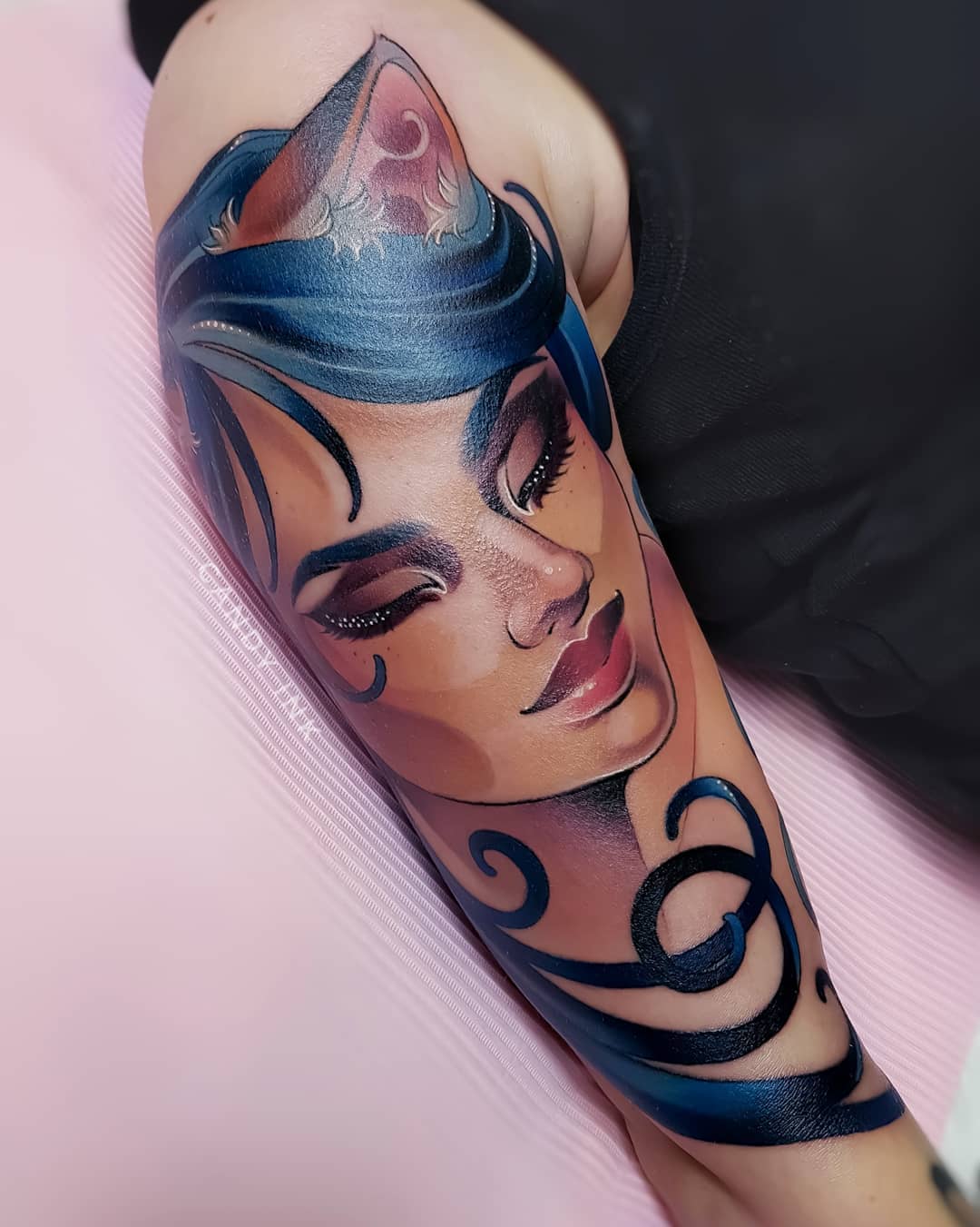 a closeup photograph showcasing a vividly colored tattoo of a woman on someone's upper arm. the tattoo is designed in a realistic style, depicting a woman with striking blue hair styled with a whimsical cat or fox ear motif. her eyes are closed, lending a serene expression to her face. the artist has expertly used shading and highlights to give the tattoo depth and dimension. the tattoo sits on the outer side of the arm, wrapping around to the back of the arm with swirling black lines. the background features a soft pink, likely the covering of the tattoo studio chair or table.