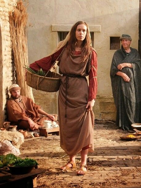 the image depicts a scene that seems to be set in ancient times, possibly biblical. a young woman, dressed in a long brown robe and sandals, is walking outdoors while carrying a basket of vegetables and herbs. she is wearing a reddishbrown overdress or shawl. another older woman in a grey robe is standing nearby, appearing to be observing her.  a man in simple clothing is seated in the background. the setting appears to be a courtyard with stone or brick buildings. the overall mood is serene and evokes a sense of historical or religious context.