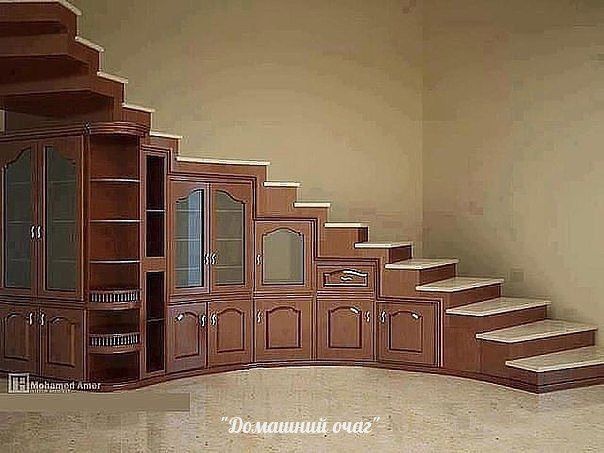 the image shows a brown wooden cabinet that transitions into a staircase. the cabinet has glass doors, shelves, and drawers, and the stairs have white tops. the cabinet and stairs are arranged in a curve. the wall behind is plain and beige.