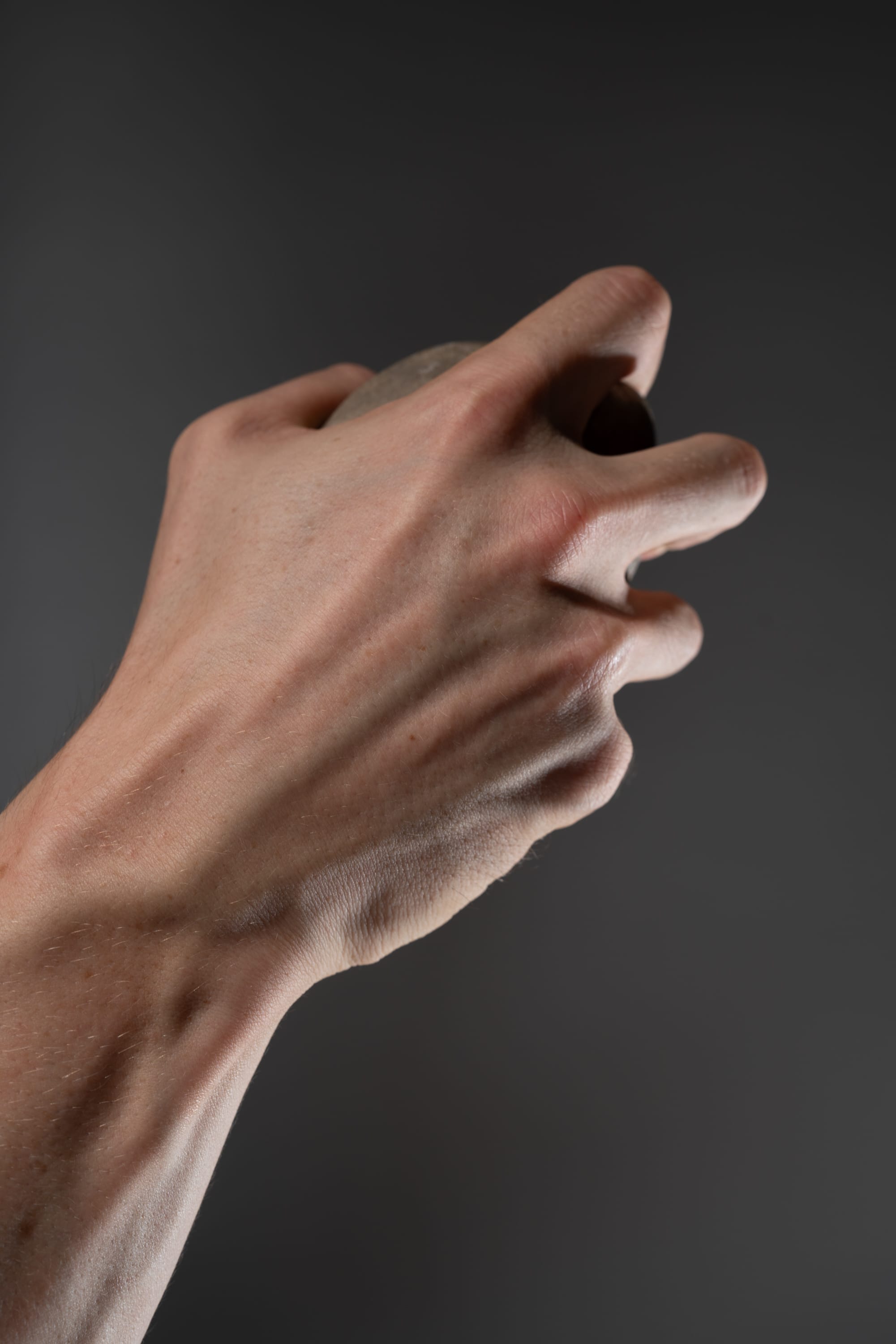 a closeup image of a fairskinned hand tightly gripping a round object that's partially concealed. the hand's veins are pronounced, showcasing the force of the grip. the background is a neutral gray, focusing attention on the hand's action and the tension in the muscles.