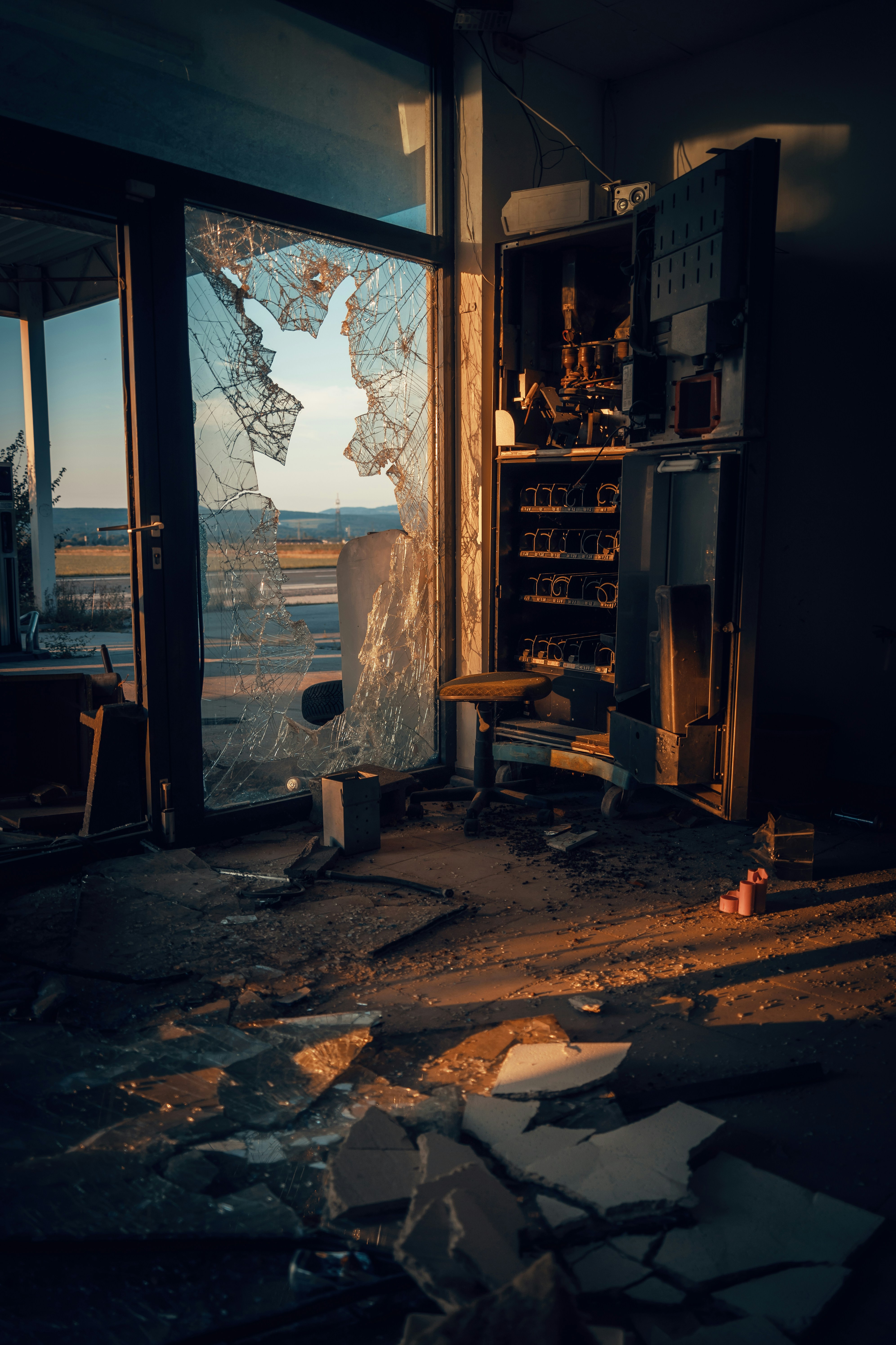 this is an image of the inside of an abandoned building. the setting sun casts an orange glow through a broken window, illuminating the debrisstrewn floor. an old vending machine stands against the wall, next to a broken chair. the scene evokes a sense of desolation and decay.