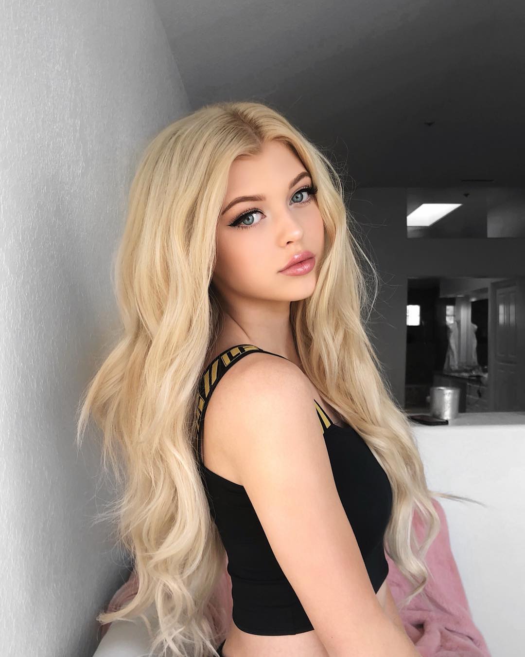 a portrait of a young woman with long blonde hair, blue eyes, fair skin and light skin. she is wearing a black tank top and looking at the camera. the background is a room with a white wall and a hallway.