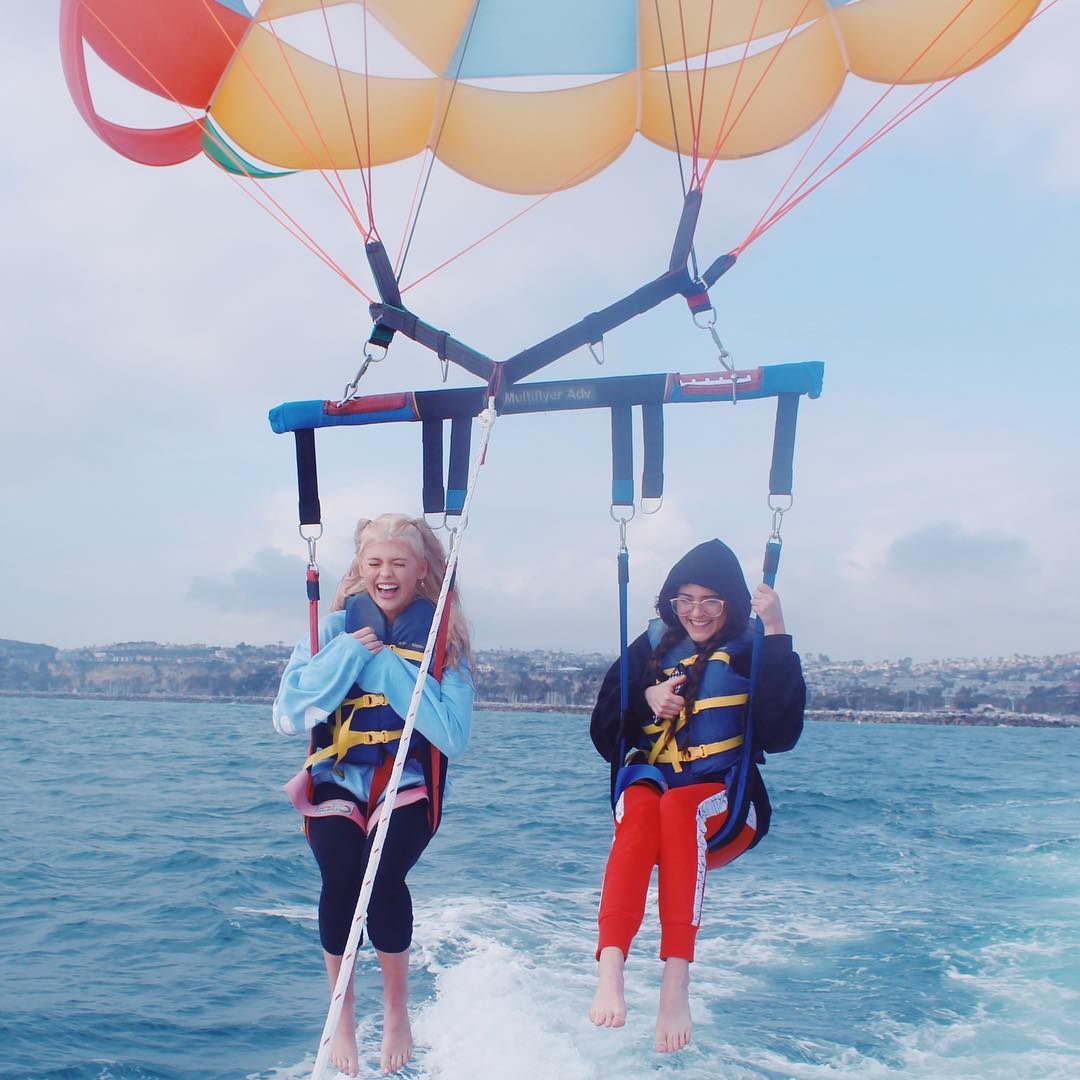 two women are parasailing over the ocean, wearing life vests and appearing to enjoy the experience. they are suspended in the air with a colorful parachute overhead. the water is blue and there is land visible in the background.