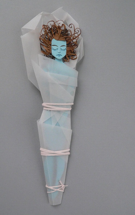 a handmade doll is wrapped in tissue paper and held together with pink bandages. the doll has blue skin and brown curly hair. it is hanging against a gray background.
