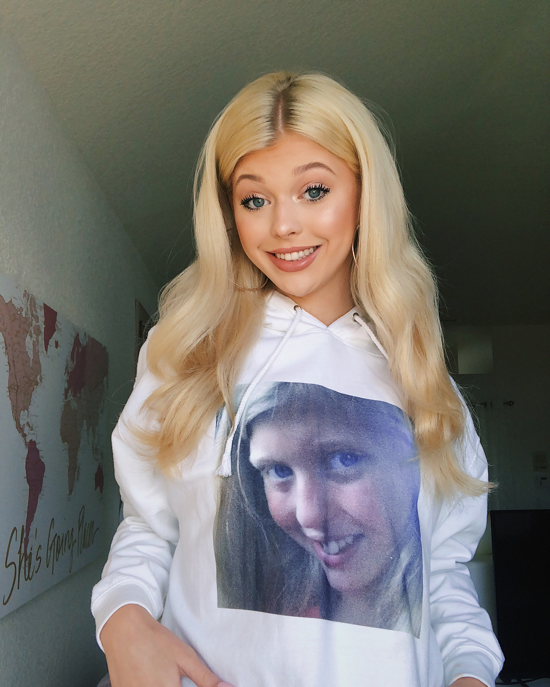 a blonde woman with blue eyes is smiling at the camera, wearing a white hoodie with a picture of a young girl on it. she has fair skin and is wearing hoop earrings. she is standing in front of a wall with a map on it.