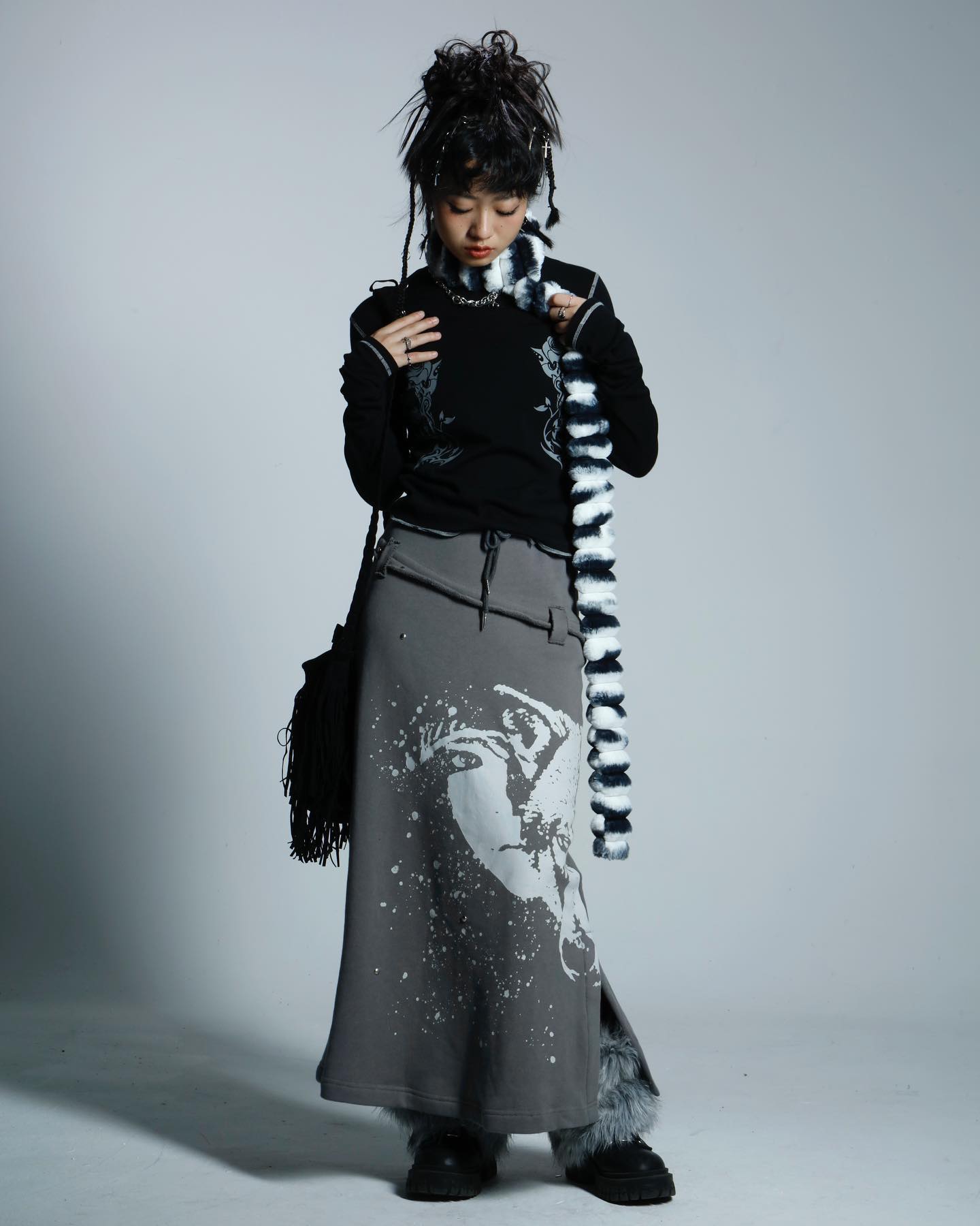 a full shot of a young woman modeling a stylish outfit. she is wearing a black longsleeve top, a grey long skirt with a graphic print, and black boots.  she's accessorized with a black and white fur scarf and a small handbag. the model's hair is styled in an edgy updo.  the backdrop is a plain grey studio setting. the overall style is alternative or grunge.