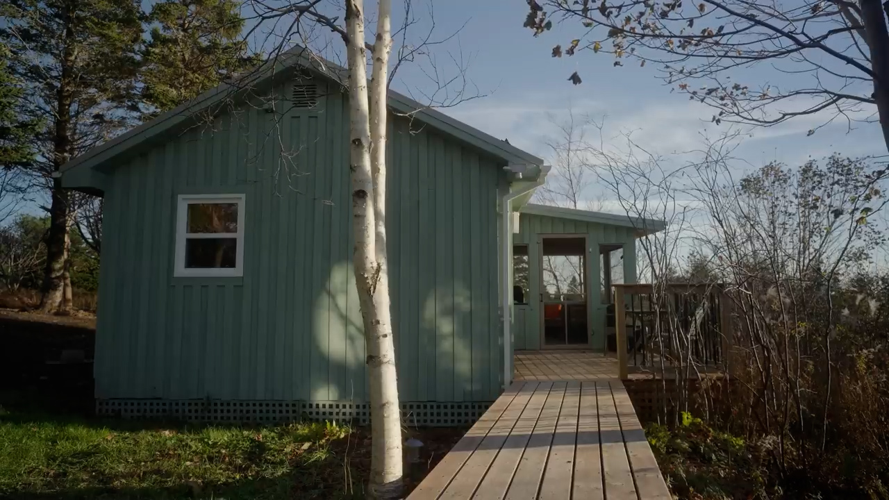 this image showcases a charming green cabin nestled amongst trees. a wooden deck extends from the side, leading to a screenedin porch. the cabin has a single window visible on this side and is bathed in warm sunlight, casting long shadows across the grass. the scene evokes a sense of tranquility and peacefulness.