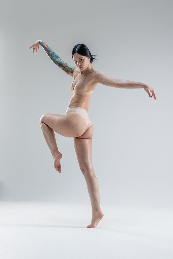 a woman with dark hair and a floral tattoo on her left arm strikes a graceful dance pose against a white background. she is wearing minimal beige underwear, emphasizing her natural body shape. the studio lighting is bright and even, highlighting her form.  she looks directly at the camera with a neutral expression.