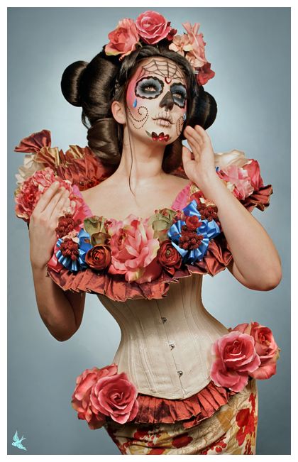 a woman poses against a muted bluegray backdrop. she is elaborately styled in dia de los muertos attire, including a painted sugar skull face, a crown of red and pink roses, and a floralembellished corset. the corset is white with red trim and accents, and features more large fabric roses. her hair is styled in a sleek updo with two buns. the overall aesthetic is dramatic and theatrical, with a focus on the artistry of the costume and makeup.