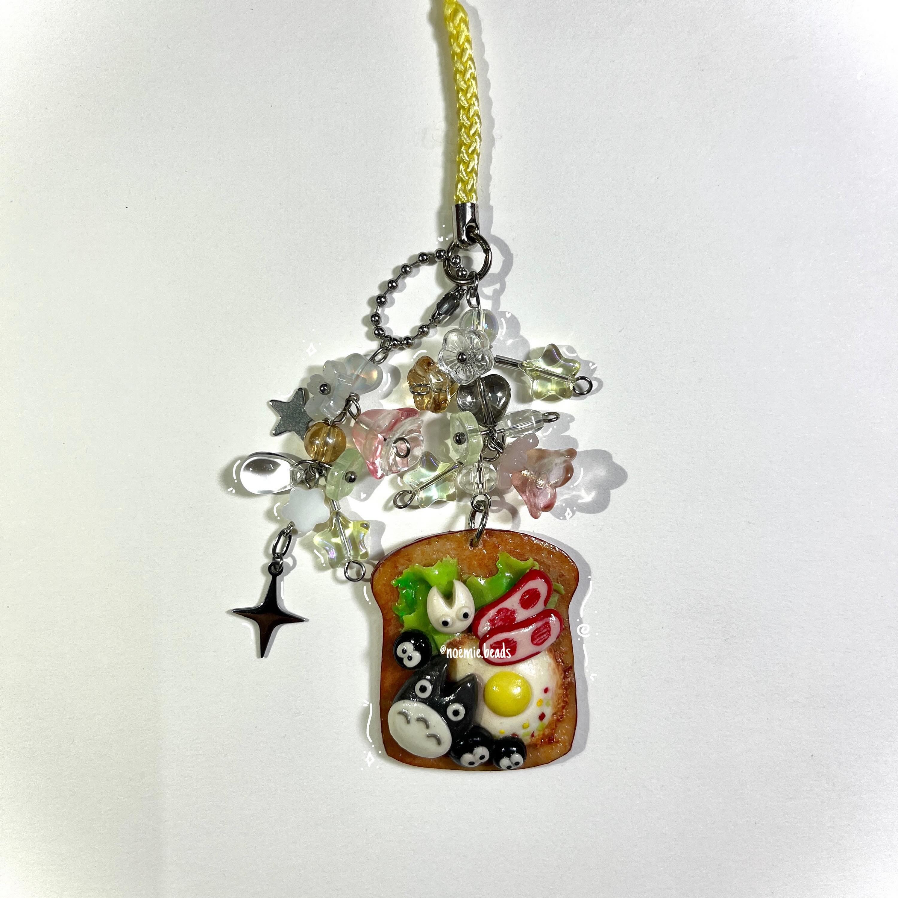 a closeup shot features a handmade charm or pendant hanging against a white background. the charm consists of a slice of toast with miniature sculptures of food items like fried egg, bacon, and lettuce, along with the characters totoro and soot sprites from the animated film "my neighbor totoro." the word "noemie beads" is visible on the toast. above the toast charm are several beads shaped like stars and flowers in colors like pink, white, and silver. a yellow cord is attached to the top of the charm, along with a chain.