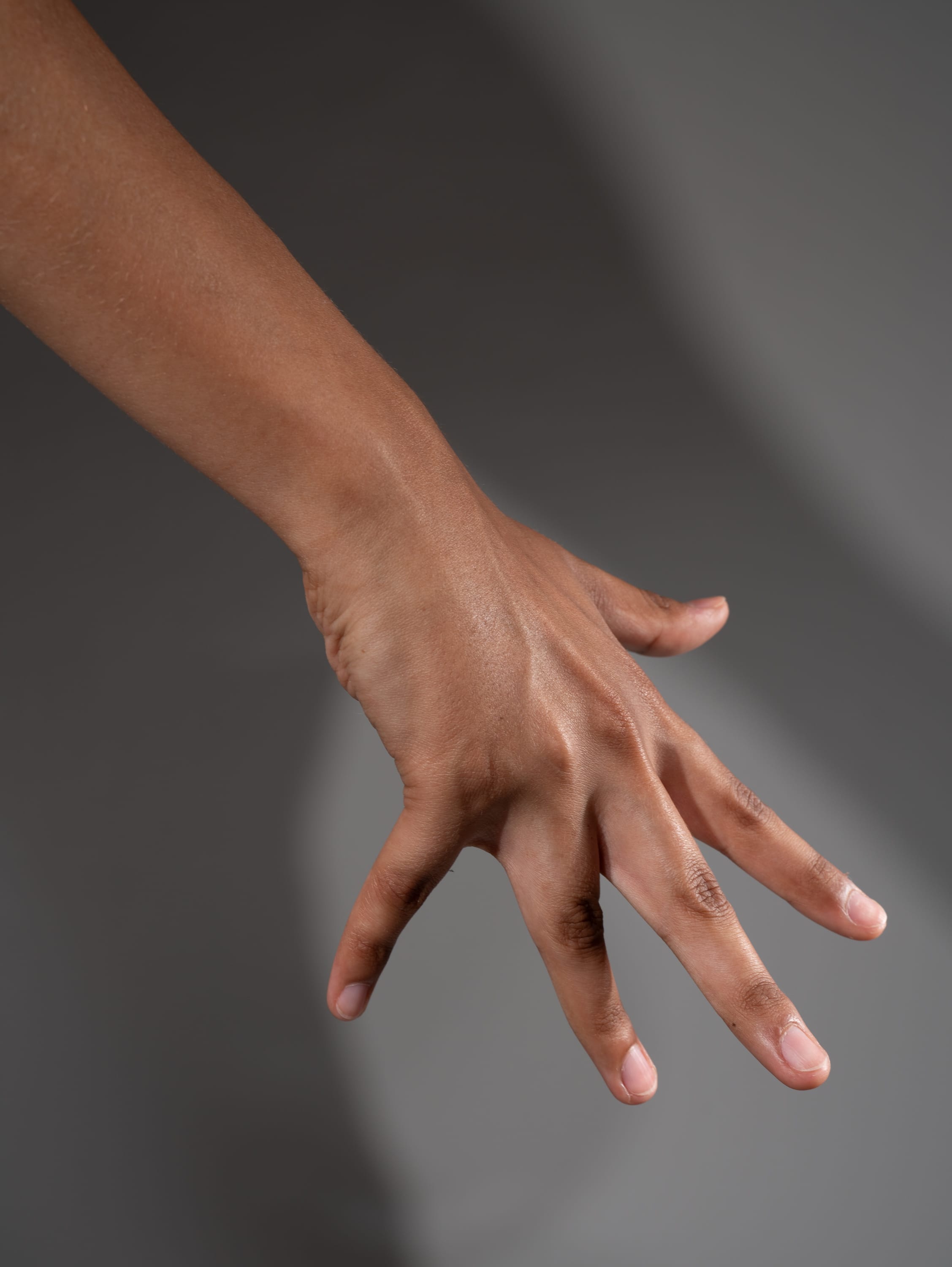 a moderately darkskinned hand reaches out from the left side of the frame, fingers outstretched against a gray background. the hand's shadow is cast onto the background.