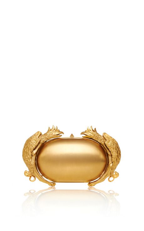 a golden clutch purse with an oblong body and an animal motif that appears to be golden eagles on either side. the whole item sits on a reflective surface.