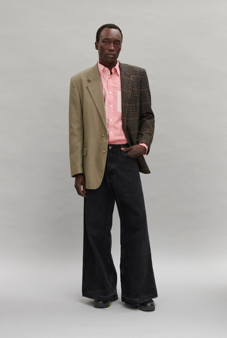 a man in a fashion studio wearing a green and brown blazer over a pink striped shirt and wide leg black jeans. he's standing in front of a gray background and looking at the camera. he is wearing black shoes.