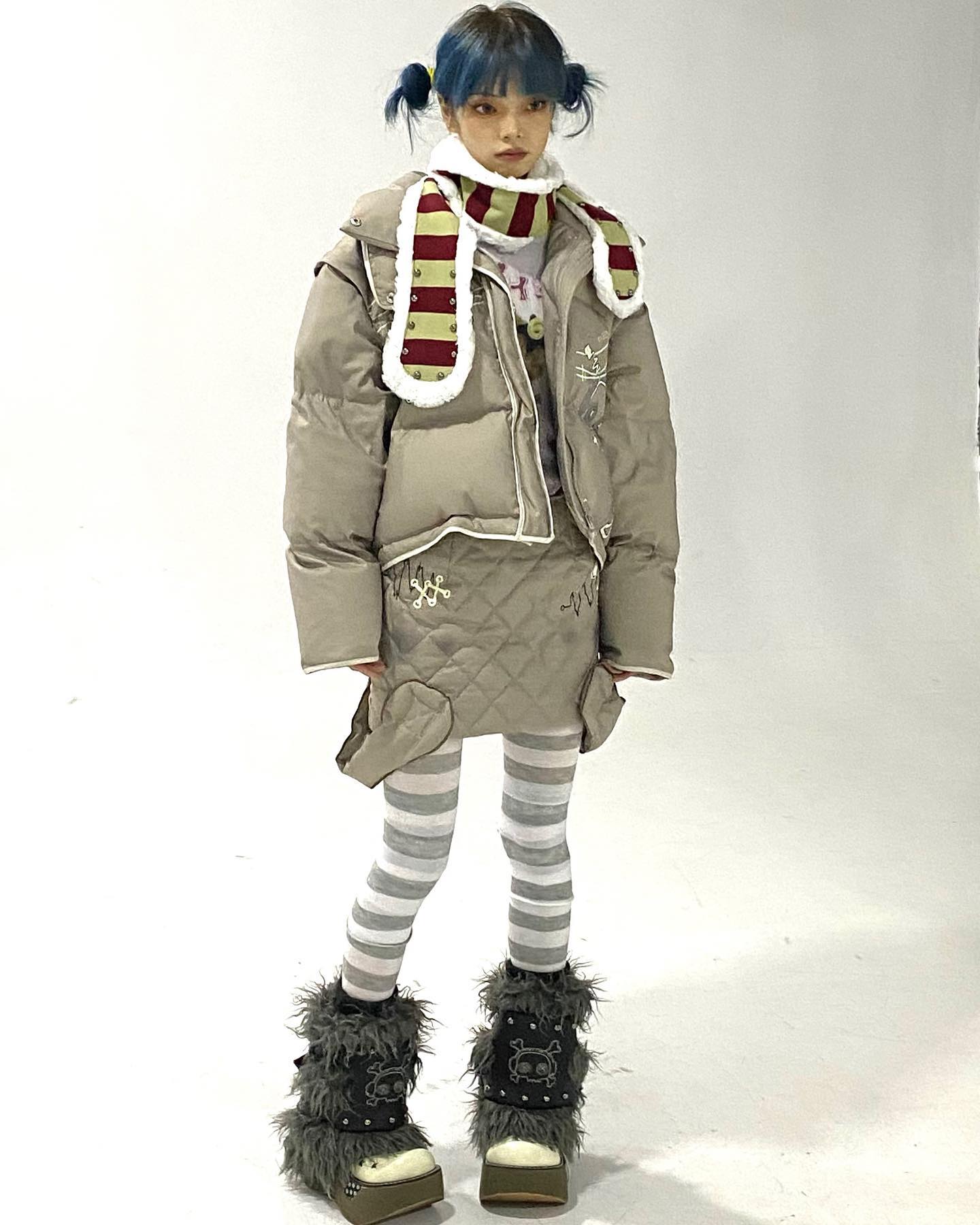 full shot of a young woman with blue hair styled in space buns, wearing a taupe puffer jacket with a white fur trim, a red and yellow striped scarf, a quilted skirt, white and grey striped tights, and furry boots with skull details. she's posing against a white background. the image is bright and welllit, showcasing the details of the outfit and accessories. the overall style is trendy and unique, blending elements of streetwear and whimsical design.