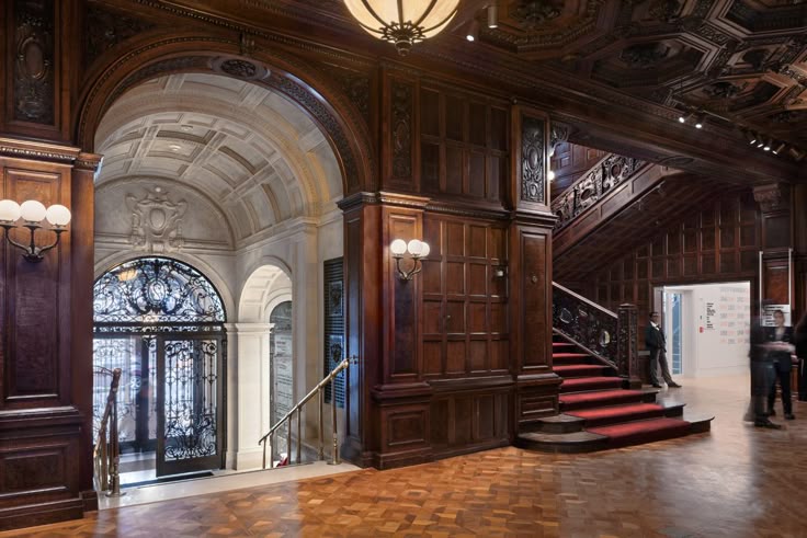 the image showcases the opulent interior of a grand mansion or historical building.  the focal point is a sweeping, redcarpeted staircase crafted from dark wood, featuring elaborate detailing and wroughtiron balustrades. the walls are adorned with rich, dark wood paneling, exhibiting intricate carvings and a high level of craftsmanship.  a large archway leads to another area of the building, displaying more ornate detailing and a wroughtiron gate.  the flooring is a polished, parquet pattern. the overall atmosphere is one of luxury, elegance, and historical significance, suggesting a setting such as a museum or private residence of high status.  ambient lighting fixtures enhance the dramatic effect.