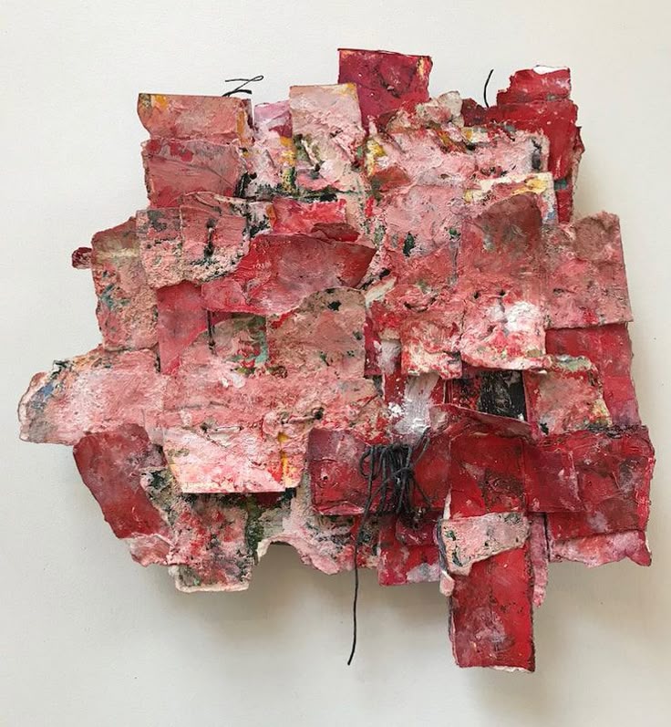an abstract mixed media piece of art is displayed against a white wall. the artwork is predominantly red and pink in color, with various geometric shapes layered and collaged together to create a threedimensional textured surface. black wire is incorporated into the design, adding to the overall texture and complexity of the piece. the artwork is an example of contemporary abstract sculpture or wall art.