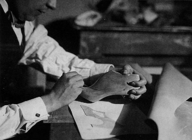 the black and white photograph shows a closeup view of a man's hands meticulously working on a wooden shoe last. he is seated and dressed in a lightcolored shirt and tie.  the image focuses on the man's hands manipulating the last, while a shoe design sketch lies on the table in front of him. the setting appears to be a workshop or studio, with indistinct background elements suggesting a historical context, possibly from the early 20th century. the overall mood is one of concentration and skilled craftsmanship.