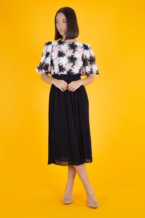 a woman is standing in front of a yellow background, wearing a cocktail dress with a floral pattern top and a black pleated skirt. she is wearing clear heels and has short brown hair.