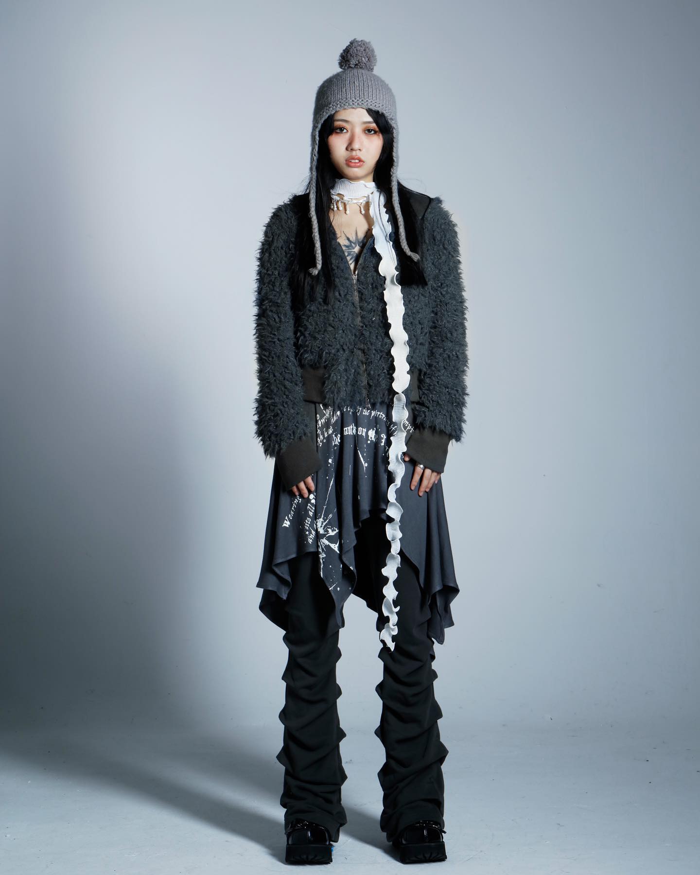 a full shot of a young woman in a fashionable outfit. she's wearing a dark gray fuzzy jacket, a ruffled gray and white skirt, stacked black pants, black boots, a gray beanie with ear flaps, and a long, thin white scarf.  the background is a plain light gray. the overall style is edgy and alternative.  the model is posing in a studio setting.